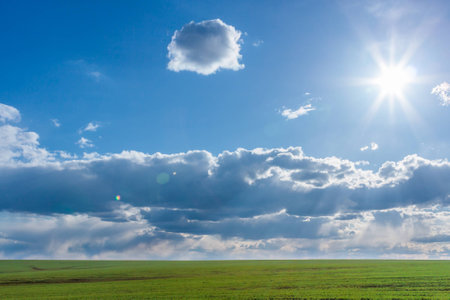 green field with fresh vibrant grass and blue sky with dramatic clouds and the sun at the daytimeの写真素材