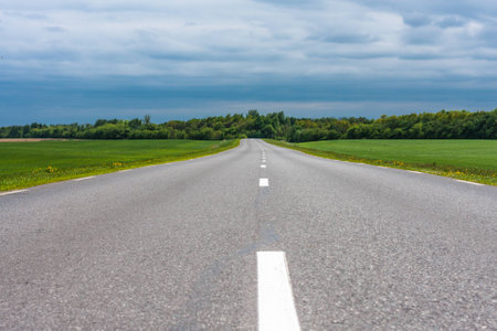 Empty asphalt country road with green grass fields under stormy clouds in the summerの写真素材