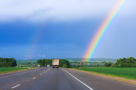 double rainbow in the blue cloudy dramatic sky over green field and a road illuminated by the sun in the country sideの写真素材