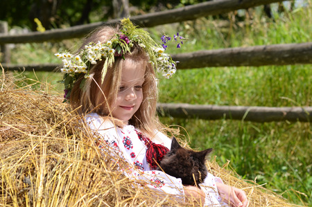 Pretty little girl in Ukrainian traditional shirt sitting on haystack with black catの写真素材