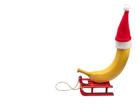 A banana in a red Santa Claus Christmas hat sits on a sled. Banana in a New Year's suit. Masquerade for a banana. Still life on a white background, isolated.の写真素材