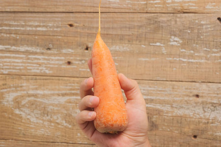 A Hand Holding a Fresh and Crisp Carrot Against a Rustic Wooden Background Surfaceの写真素材