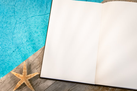 Open book without inscription seen from above on a wooden paving above a swimming pool with a starfish. Holiday atmosphere in summer.の写真素材