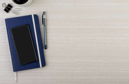 Note book with pen, smart phone, coffee mug and paper clips over wooden desk with room for text. Top...