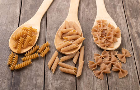 Assorted whole wheat pasta on spoons over wooden table.の写真素材