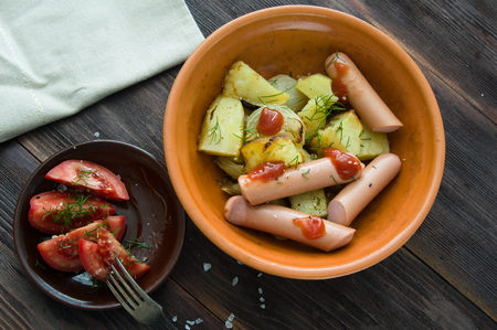 Boiled sausages with a side dish of baked potatoes and fresh sliced tomatoes on a wooden tableの写真素材