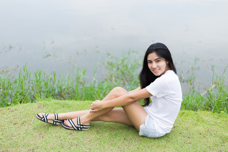 Young Asian woman is sitting on the green grass near the lakeの写真素材