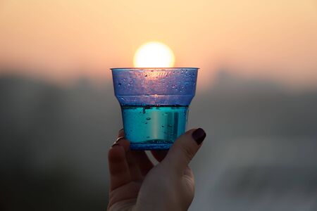 Woman's hand holding glass against setting sun with blurry skyscrapers in cityscape background.の写真素材