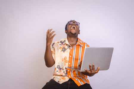 Excited young black African male holding a laptop, raising up his hands in excitement after winning...