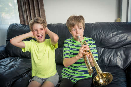 Two kids sitting on a sofa.
One is playing a trumpet and the other child annoyed is covering his ears with his handsの写真素材