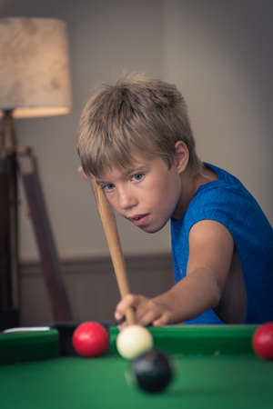 Cute boy in blue t shirt plays billiard or pool in club. Young Kid learns to play snooker. Boy with billiard cue strikes the ball on table. Active Leisure, sport, hobby conceptの写真素材