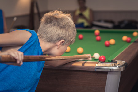 Cute boy in blue t shirt plays billiard or pool in club. Young Kid learns to play snooker. Boy with billiard cue strikes the ball on table. Active Leisure, sport, hobby conceptの写真素材