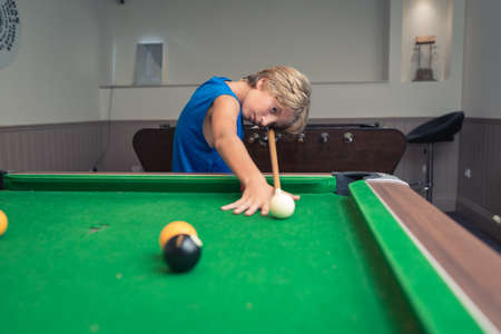 Cute boy in blue t shirt plays billiard or pool in club. Young Kid learns to play snooker. Boy with billiard cue strikes the ball on table. Active Leisure, sport, hobby conceptの写真素材