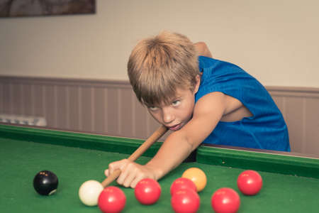 Cute boy in blue t shirt plays billiard or pool in club. Young Kid learns to play snooker. Boy with billiard cue strikes the ball on table. Active Leisure, sport, hobby conceptの写真素材
