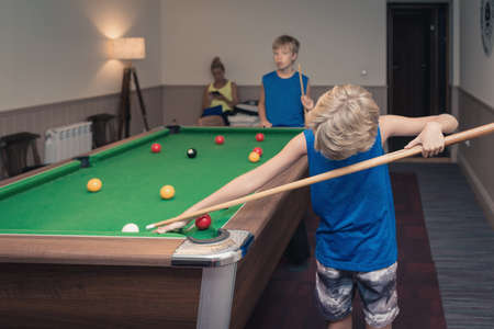Cute boy in blue t shirt plays billiard or pool in club. Young Kid learns to play snooker. Boy with billiard cue strikes the ball on table. Active Leisure, sport, hobby conceptの写真素材