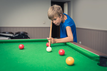 Cute boy in blue t shirt plays billiard or pool in club. Young Kid learns to play snooker. Boy with billiard cue strikes the ball on table. Active Leisure, sport, hobby conceptの写真素材