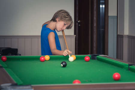 Cute boy in blue t shirt plays billiard or pool in club. Young Kid learns to play snooker. Boy with billiard cue strikes the ball on table. Active Leisure, sport, hobby conceptの写真素材