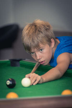 Cute boy in blue t shirt plays billiard or pool in club. Young Kid learns to play snooker. Boy with billiard cue strikes the ball on table. Active Leisure, sport, hobby conceptの写真素材