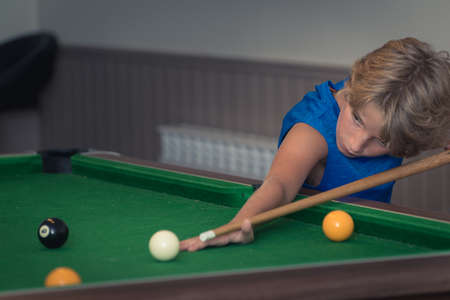 Cute boy in blue t shirt plays billiard or pool in club. Young Kid learns to play snooker. Boy with billiard cue strikes the ball on table. Active Leisure, sport, hobby conceptの写真素材