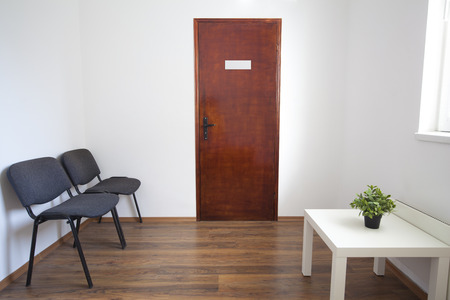Small white waiting room without people. A doctor, dentist or other medical practitioner provides this room for the use of people who are waiting to be seen. The wooden door is closed and furniture is two chairs, small table with green plant.の写真素材