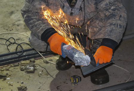 The hands of the worker in gloves cut metal profile with a grinding machine. orange sparks fly from...