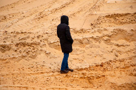 A tall slim man from his back in a black elongated jacket with a hood on his head walks in the sand in a quarry between the sandy holly and dunes, with trails from tractors and cars.の写真素材