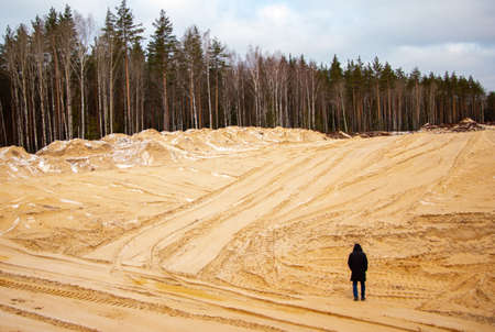 A tall slim man from his back in a black elongated jacket with a hood on his head walks in the sand in a quarry between the sandy holly and dunes, with trails from tractors and cars.の写真素材