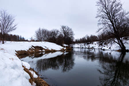 A forest river with a fast flow and small waves flows between snowy shores with trees and bushes...