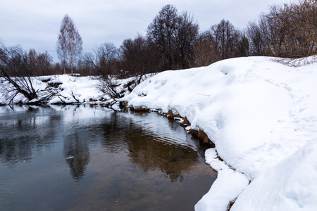 A forest river with a fast flow and small waves flows between snowy shores with trees and bushes...
