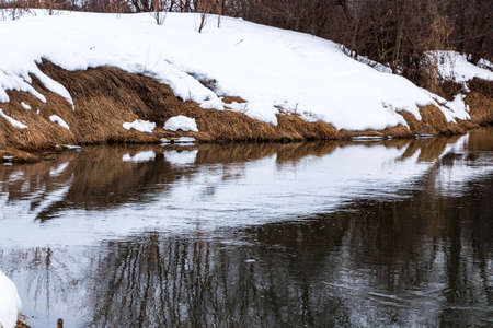 A forest river with a fast flow and small waves flows between snowy shores with trees and bushes...