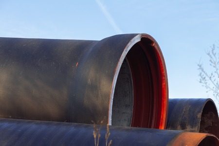 Large diameter metal pipes for water supply lie on storage areas under a blue sky