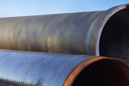 Large diameter metal pipes for water supply lie on storage areas under a blue sky