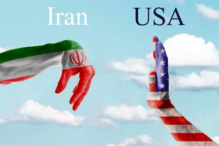 The concept of two hands of mature men painted in the colors of the flags of Iran and the United States against a blue sky. Military confrontation between the two nuclear powers.の写真素材