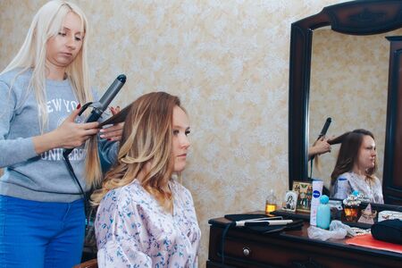 August 10, 2019 Minsk Belarus The master of hairstyles puts hair in the girl in front of the mirror with the help of hair tongsのeditorial素材