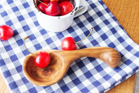 Composition of cup, wooden spoon and berries. Cherries is the reason of the longevity. White retro cup, wooden spoon and berries on a checkered napkin on the table. Wooden handmade spoon with retro cupの写真素材