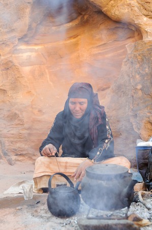 WADI MUSA, JORDAN - NOVEMBER 18, 2012: Old woman cooking tea for tourist on desert sands of ancient Petra city. Petra is historical and archaeological city and famous for its rock-cut architecture and water conduit system.Another name for Petra is the Rosのeditorial素材