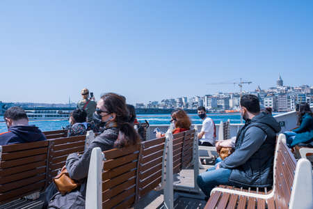 Istanbul, Turkey - 04.29.2021: several Turkish ship passengers with protective masks sit on seats and look at sea and Galata Bridge and Tower while travelling along Marmara Sea in a clear air with copy spaceのeditorial素材