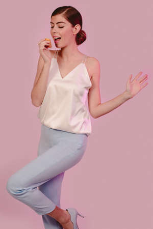 Cheerful pretty stylish young girl fooling around and eating in yellow and pink dress looking at camera. Isolated on pink background. Design copy space.の写真素材