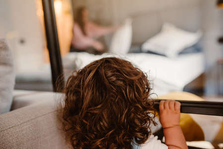 Adorable toddler with long curly hair looking through a window of a room at his motherの写真素材