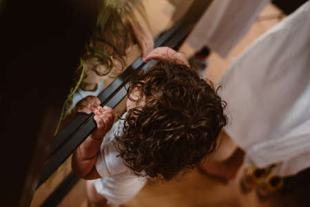 Unrecognizable toddler with long curly hair looking through a window of a room at his motherの写真素材