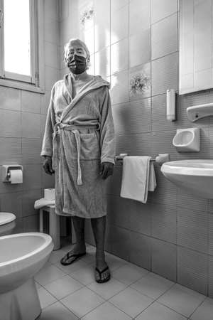 Middle-aged man in the bathroom, wearing a white bathrobe and protective maskの写真素材