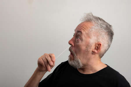 middle aged man with gray hair and beard using the coronavirus (covid-19) antigen rapid self-test kit (atk) at home with a cotton nasal swabの写真素材