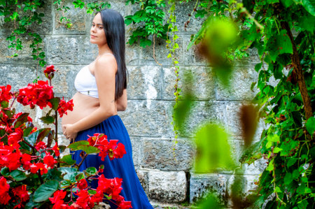 Side view of a pregnant woman standing in a lush garden beside a stone wall. She wears a white top...