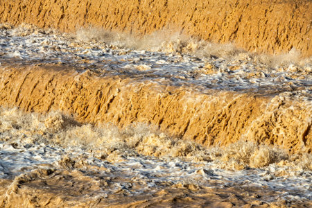 Dynamic close-up image of a cascading waterfall filled with muddy water, captured during a flood season. The strong current and thick sediment create a striking visual of raw natural power and chaos. The turbulence and foam in the water suggests heavy rainfall or upstream erosion. This image highlights the environmental effects of extreme weather and flooding, making it suitable for use in environmental awareness campaigns, news articles, climate change studies, and educational materials. The texture and color of the water add a dramatic and almost abstract quality to the composition, emphasizing both the beauty and destructive potential of nature.の写真素材