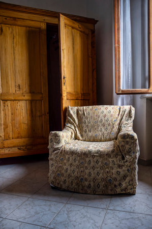 a cozy rustic interior featuring an upholstered armchair with a patterned fabric cover placed beside a large wooden wardrobe. The wardrobe, crafted from natural wood, has one door partially open, adding depth and atmosphere to the composition. Sunlight filters through an open window, softly illuminating the scene and highlighting the textures of the furniture. The room conveys a warm, vintage, and homely atmosphere, typical of traditional Italian interiors.の写真素材