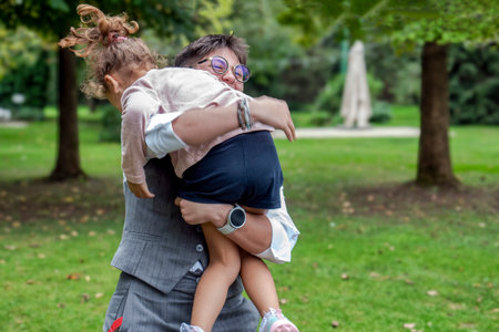 A joyful adult holding and hugging a young child in a green park. The image conveys love, care, family bonding, and emotional connection in an outdoor natural environment. Ideal for concepts of parenting, childhood, happiness, trust, and everyday family life.の写真素材