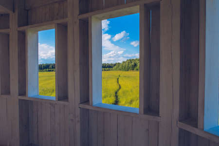 Path in the grass field through the wooden window view. Meadow picturesque summer landscape with clouds on blue sky background. Window portal to the nature landscape.の写真素材