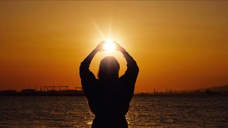 A high quality silhouette photograph showing hands framing the sun at golden hour during a dramatic...