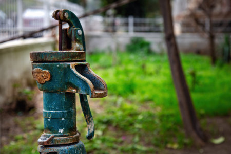 Old rusty green manual water pump in a lush garden background. This vintage cast iron hand pump features weathered textures and oxidation, symbolizing traditional farming, rural life, and historical irrigation methods.