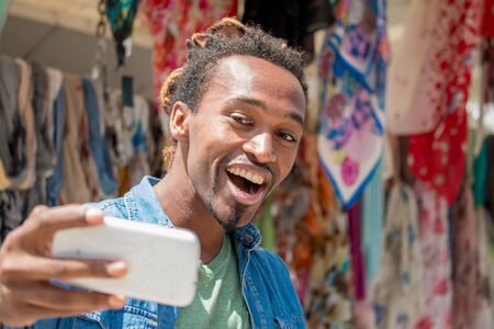 happy african guy smiling and taking a selfie photo using smartphone. cheerful man looking at the phone with a wide toothy smile on a bright sunny dayの写真素材